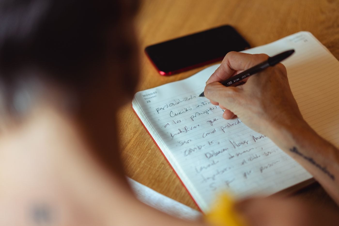 A writer adds handwritten notes to an open notebook at a wooden table.