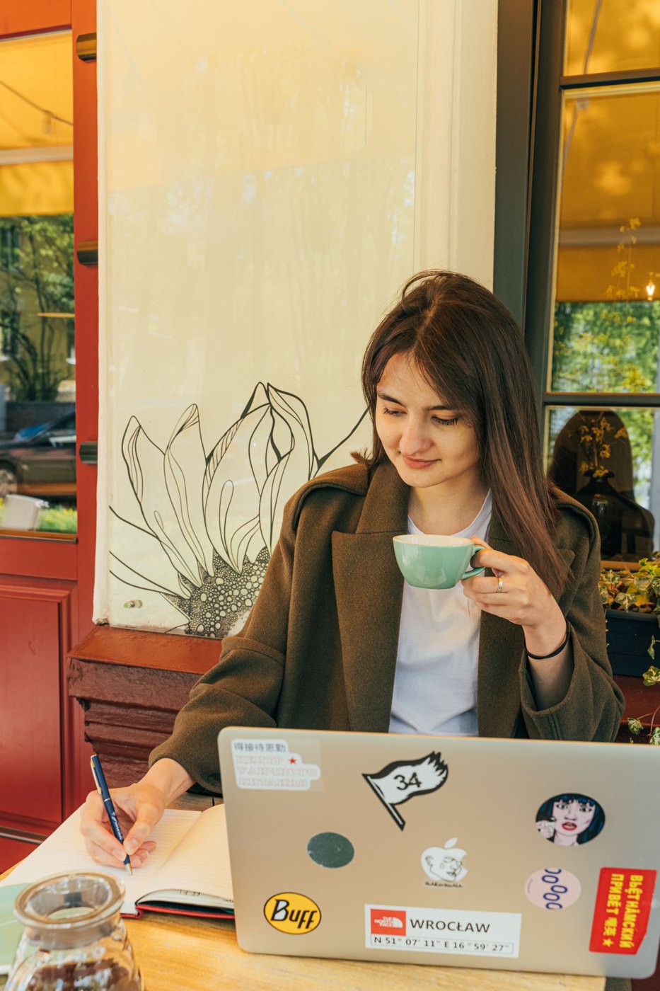 A writer works in a notebook beside a laptop at a cafe.
