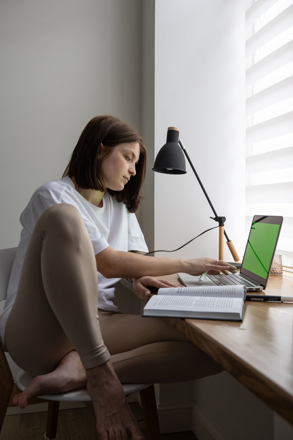 A reader using a book beside an open laptop at a desk.